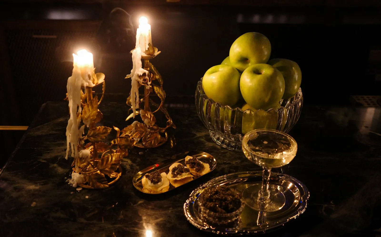 Candles, green apples, a coupe and small bites arranged on a dark table at Dandelion